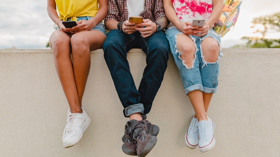 Legs in sneakers of young company of friends sitting park using smartphones, man and women having fun together, colorful summer hipster fashion style, communication wireless connecting devices